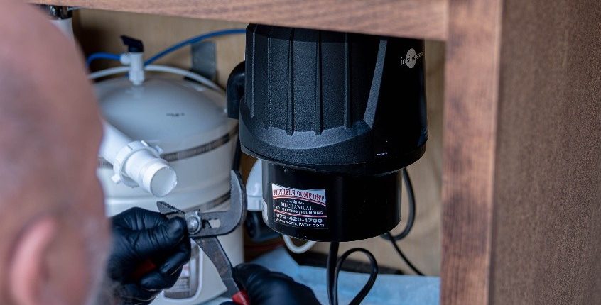 Plumbing technician using a wrench to work on a garbage disposal beneath a kitchen sink, featuring Southern Comfort Mechanical branding.