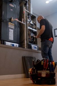 Professional HVAC technician performing maintenance on an indoor furnace and air handler system with tools and panels removed.