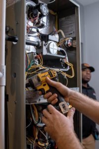 Professional HVAC technician servicing a residential furnace, using a power drill to access internal components while another technician observes.